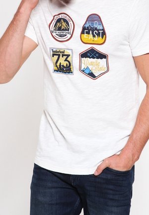 Biała koszulka z czterema kolorowymi naszywkami przedstawiającymi góry oraz napisy: „New Hampshire”, „North East”, „Timberland 73” i „Granite State”, noszona przez mężczyznę.