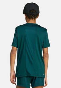 Kind in een donkergroene sportshirt en -short, met een zwarte pet, van achteren gezien tegen een effen lichte achtergrond.