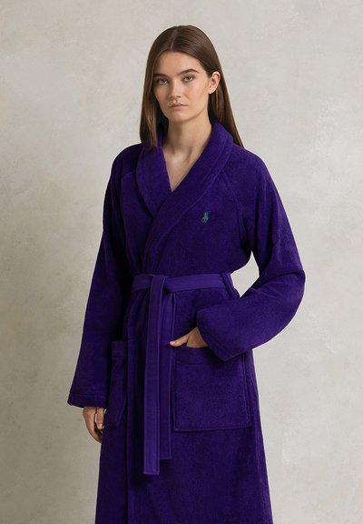 Peignoir de bain en éponge violet avec col châle, ceinture et deux poches frontales. Présente un petit logo vert sur la poitrine. Texture douce.