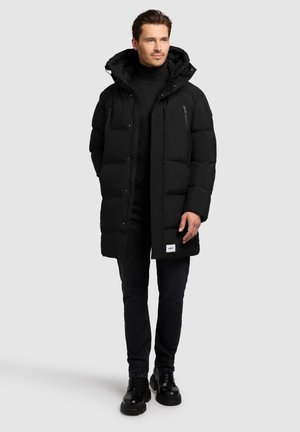 Schwarze Puffjacke mit hohem Kragen, gestepptem Design und einem Markenlabel am Saum, kombiniert mit dunklen Jeans und schwarzen Schnürstiefeln.