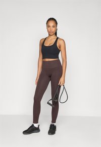 Fekete crop top, bordás, magas derekú, sötétbarna leggingszel párosítva. A modell fekete ellenállási szalagot tart, fekete sportcipőben.