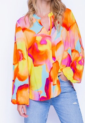 Mujer vistiendo una blusa con un patrón abstracto y colorido en tonos naranja, amarillo, rosa y azul, y unos vaqueros azules claros rasgados.