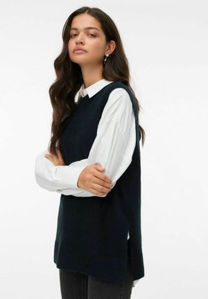 Pull sans manches en tricot bleu marine superposé à une chemise blanche à boutons, avec une coupe décontractée et des fentes sur les côtés. Design simple sans motifs.