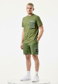 Oliven grønn kortermet skjorte med en lomme på brystet og matchende shorts. Har glidelåsdetalj og et svart logo-merke. Båret med hvite sneakers.