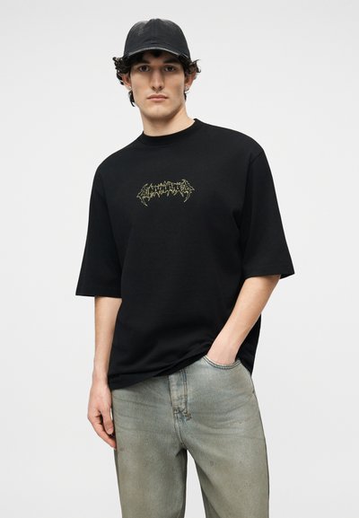 Νέος άνδρας που φοράει μαύρο καπέλο, φαρδιά μαύρη μπλούζα με γραφικά και ξεθωριασμένο ανοιχτό μπλε τζιν, στέκεται με το ένα χέρι στην τσέπη.