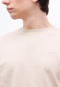 Νεαρός που φοράει μπεζ T-shirt Levi's με στρογγυλή λαιμόκοψη, στραμμένος προς τα αριστερά με το κεφάλι ελαφρώς γυρισμένο, ορατός ο λαιμός και μερικό πρόσωπο.