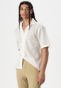 Chemise blanche à manches courtes avec un motif en zigzag, fabriquée en tissu tricoté transparent, dotée d'un col plat et d'une fermeture à boutons. Coupe décontractée.
