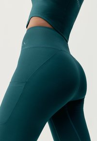 Turkusowe legginsy sportowe z wysokim stanem i dopasowanym krojem. Gładka tekstura i szczegół kieszeni z tyłu. Pionowe szwy na bokach.