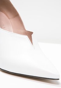 Chaussure blanche en cuir à bout pointu avec une découpure en V. Texture lisse, couture minimale et un léger talon. L'intérieur est d'un beige doux.