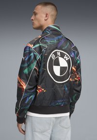 Svart vadderad bomberjacka med färgglada svängor, med en framträdande vit BMW-logotyp på baksidan och ribbade mudd och fåll.