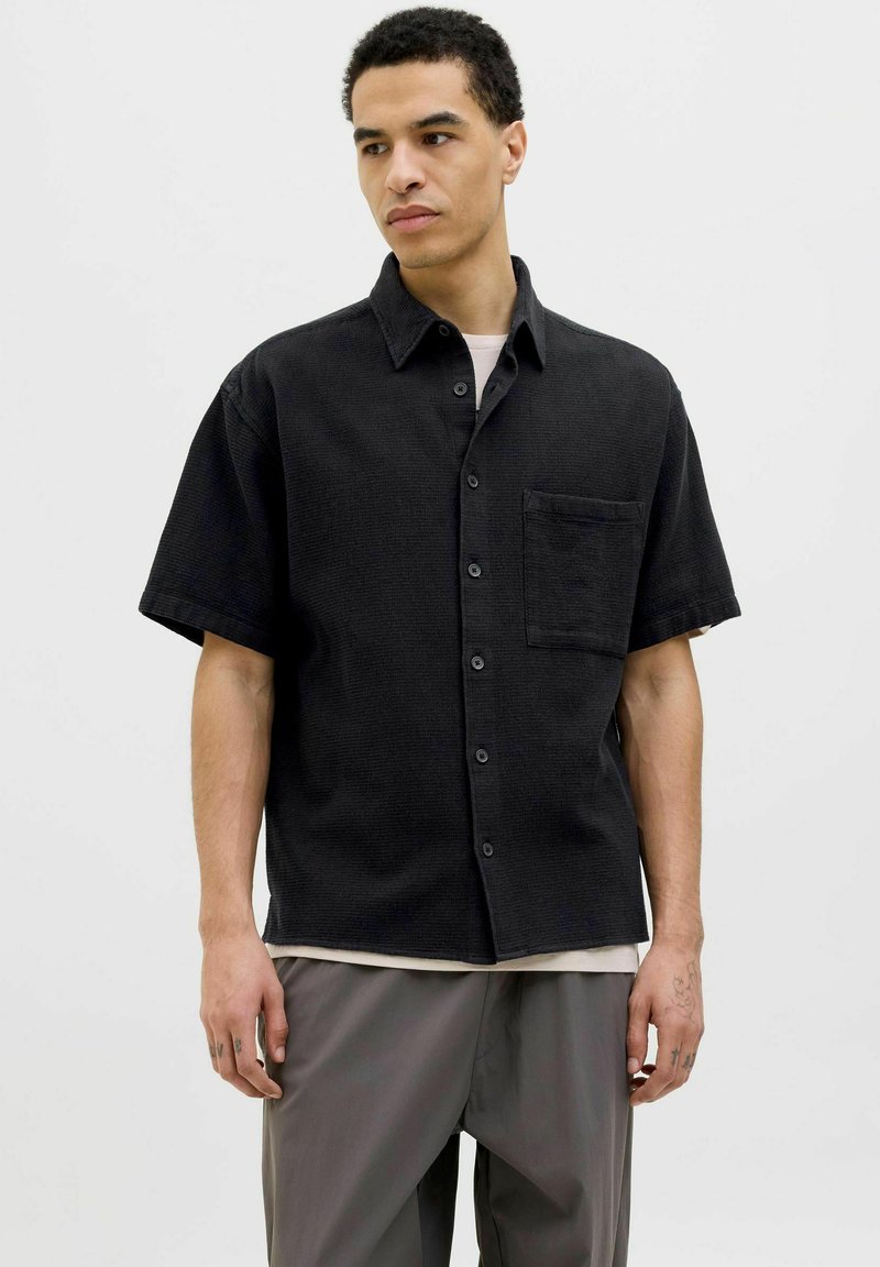 Chemise noire à manches courtes avec un tissu texturé, poche frontale unique et col, portée par-dessus un sous-vêtement de couleur claire.