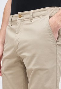 Μπεζ casual παντελόνι με κούμπωμα κουμπιού και πλαϊνές τσέπες, ένα χέρι εν μέρει μέσα στην τσέπη, συνδυασμένο με μαύρο πουκάμισο.