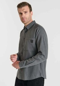 Camisa de manga larga gris de tejido suave con cierre de botones en la parte delantera, un bolsillo en el pecho izquierdo y un sutil detalle de logo.