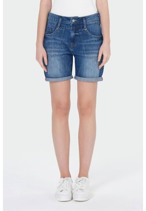 Femme portant un short en denim bleu roulé et des baskets blanches, debout devant un fond clair uni.