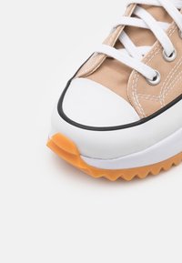 Zbliżenie beżowego sportowego obuwia z białą gumową osłoną palców, białymi sznurowadłami, czarnymi wykończeniami i pomarańczową ząbkowaną podeszwą na białym tle.
