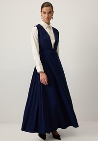 Rochie maxi bleumarin cu un fusta plisata, decolteu adânc în V și un corset mulat. Purtați-o peste o cămașă albă cu guler. Textură de țesătură elegantă.
