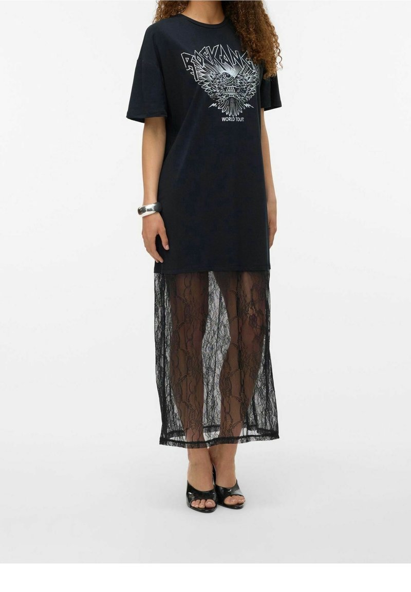 Nainen pukeutuneena mustaan graafiseen T-paita-mekkoon, jossa on "Rock Angel" -painatus ja läpikuultava mustasta pitsistä valmistettu hame, yhdistettynä mustiin avointoisiin korkokenkiin.