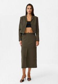 Blazer court olive avec trois boutons, accompagné d'une jupe crayon longue assortie avec une fente à l'avant. Des escarpins à brides noires complètent le look.