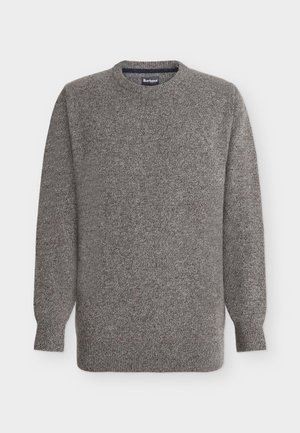 Jeune homme debout portant un pantalon beige, des baskets beiges, une chemise blanche et un pull gris, regardant légèrement sur le côté les mains dans les poches.