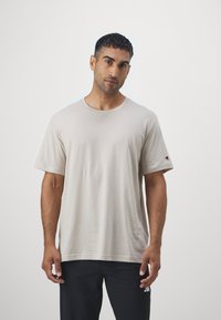 Beige kortermet t-skjorte laget av mykt stoff, med rund hals og et lite logo-patch på venstre erme. Enkel design.