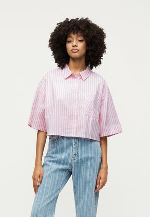 Frau mit lockigem Haar, die ein locker sitzendes, kurzärmeliges pink-weiß gestreiftes Shirt und hoch taillierte blaue Jeans mit vertikalen Streifen trägt.