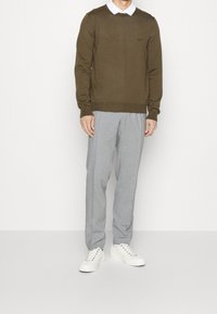 Brun långärmad tröja över en vit skjorta med krage, parat med grå skräddarsydda byxor och vita sneakers med flerfärgade ögondroppar.