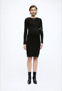 Σκούρο πράσινο πλεκτό φόρεμα με ριγέ υφή, μακρυά μανίκια, εφαρμοστή γραμμή, στρογγυλή λαιμόκοψη και μήκος μέχρι τη μέση της γάμπας. Συνδυασμένο με μαύρα μποτάκια αστραγάλου και μικρό τσαντάκι.