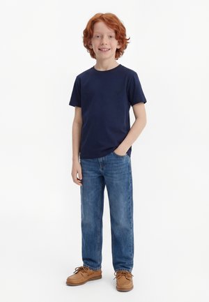 Roodharige jongen die met één hand in zijn zak staat, gekleed in een marineblauw T-shirt, blauwe spijkerbroek en bruine veterschoenen, tegen een witte achtergrond.