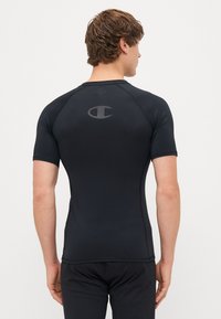 T-shirt de sport noir à manches courtes en tissu lisse et ajusté. Présente un logo gris dans le haut du dos et un design de manches raglan.