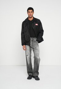 Μαύρο μπουφάν από νάιλον με το λογότυπο "DIESEL", φορεμένο πάνω από ένα μαύρο πουκάμισο. Γκρι distressed τζιν με σκισίματα. Μαύρα αθλητικά παπούτσια. Απλός, μοντέρνος σχεδιασμός.