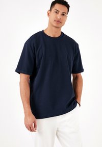 Tricou bleumarin cu mânecă scurtă și guler rotund, cu un material texturat și manșete răsucite. Purtați-l cu pantaloni albi.