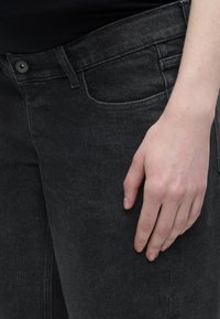 Το δεξί χέρι ακουμπάει στο πάνω μέρος του μηρού, φορώντας σκούρα γκρι τζιν με ορατή μπροστινή τσέπη και κούμπωμα με κουμπί.