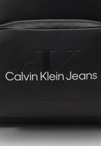 Borsa in pelle nera con logo in rilievo 'CK' e testo 'Calvin Klein Jeans' in bianco. Include un dettaglio con zip argentata nella parte superiore.