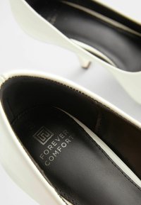Talons hauts en cuir verni blanc avec un bout pointu, dotés d'une doublure intérieure noir et d'un petit talon. Logo "Forever Comfort" en relief à l'intérieur.
