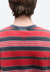 Κοντά καστανά μαλλιά σε άτομο που φοράει κόκκινο και γκρι ριγωτό πλεκτό πουλόβερ, φωτογραφημένο από πίσω, με φόντο έναν απλό φωτεινό τοίχο.