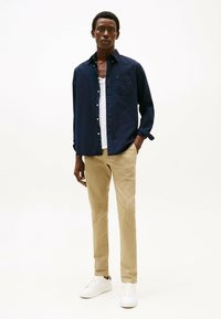 Marineknopen overhemd, lichte tanktop eronder, beige chino's en witte sneakers. Het overhemd heeft een borstzak en opgerolde mouwen.