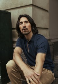 Laivastonsininen poolopaita, jossa on ruskeat yksityiskohdat, lyhyet hihat ja kolme nappia; yhdistetty vaaleanruskeisiin chinoihin; poseeraa neutraalia taustaa vasten.