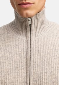 Beżowy sweter z prążkowanego dzianiny z wysokim kołnierzem, z metalowym suwakiem. Zbliżenie podkreśla teksturę i detale projektu.