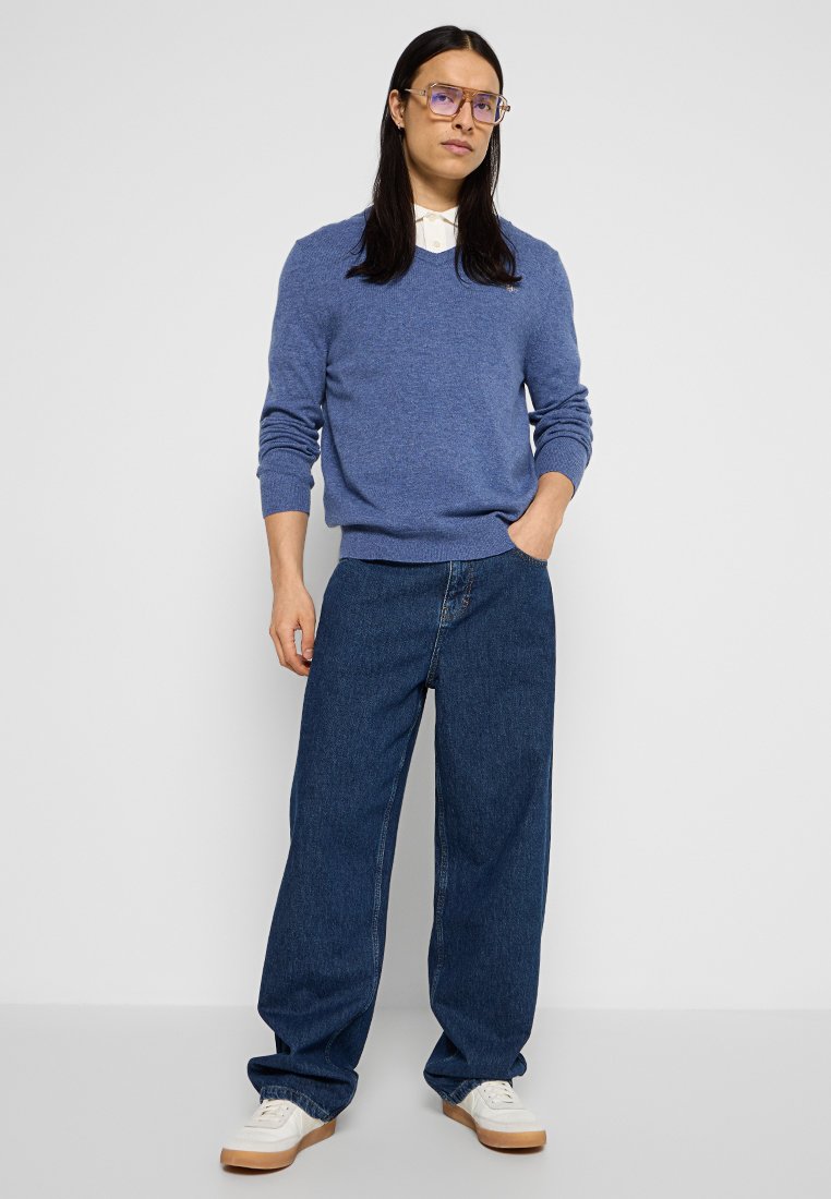 Niebieski sweter dzianinowy z dekoltem w kształcie litery V, w połączeniu z luźnymi ciemnymi dżinsami i białymi trampkami. Okulary z jasną oprawką i przezroczystymi soczewkami.