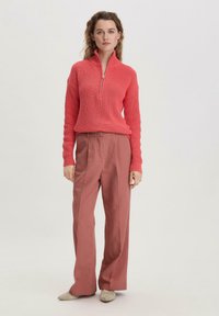 Korallenfarbener, strukturierter Pullover mit Reißverschluss und weit geschnittenen, hoch taillierten, pinken Hose. Modell steht frontal. Neutraler Hintergrund.