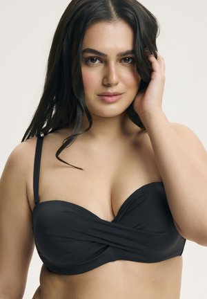 Mladá žena s dlhými tmavými vlasmi, ktorá má na sebe čierny bikiny top, dotýka sa vlasov a pozerá sa priamo do kamery s neutrálnym výrazom.