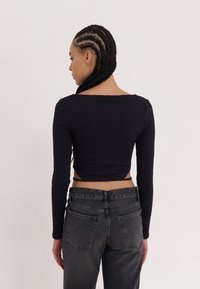 Black long-sleeve cropped top with a textured pattern, paired with high-waisted dark gray jeans. Visible back design with ties at the waist.