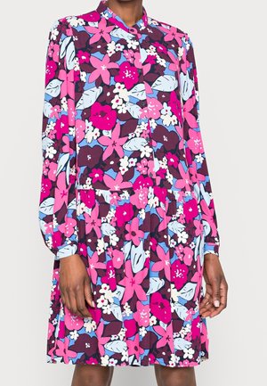 Žena má na sobě dlouhý halenkový šaty s knoflíky a výrazným květinovým vzorem v růžových, fialových, modrých a bílých barvách.