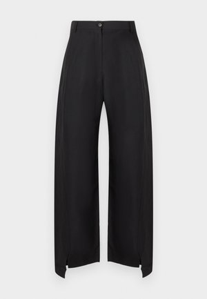 Femeie cu păr scurt purtând o cămașă palid galbenă cu nasturi peste un maiou alb, pantaloni negri largi și pantofi negri, stând cu o mână în buzunar.