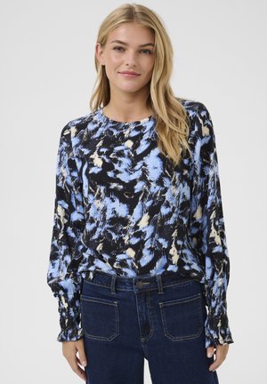Blouse à manches longues en imprimé abstrait bleu et noir, avec encolure ronde et poignets froncés. Associée à un jean bleu foncé.