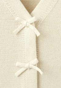 Primo piano della parte frontale di un cardigan lavorato a maglia color crema con due chiusure a fermaglio in tessuto abbinato, legate a fiocco.