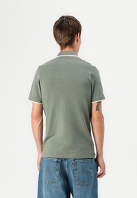 Groene poloshirt met korte mouwen, voorzien van een kraag en witte afwerking. Draagt met blauwe spijkerbroek, achteraanzicht weergegeven.