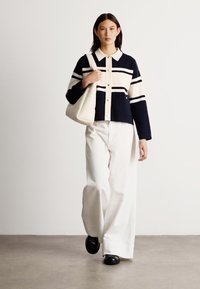 Tamsiai mėlynos ir grietinėlės spalvos dryžuotas megztinis su apykakle, derinamas su plačiomis baltomis kelnėmis ir juodais batais. Modelis laiko grietinėlės spalvos rankinę.