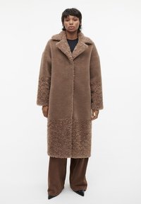 Bruine, lange jas gemaakt van zachte, gestructureerde stof met een gesplitst ontwerp. Heeft een brede kraag en opvallende mouwen. Draagbaar met zichtbare bruine broek.