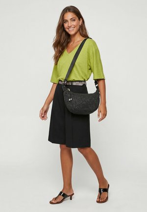 Junge Frau lächelt, trägt ein limettengrünes Top, schwarzen knielangen Rock, schwarze Sandalen und trägt eine schwarze geflochtene Umhängetasche.